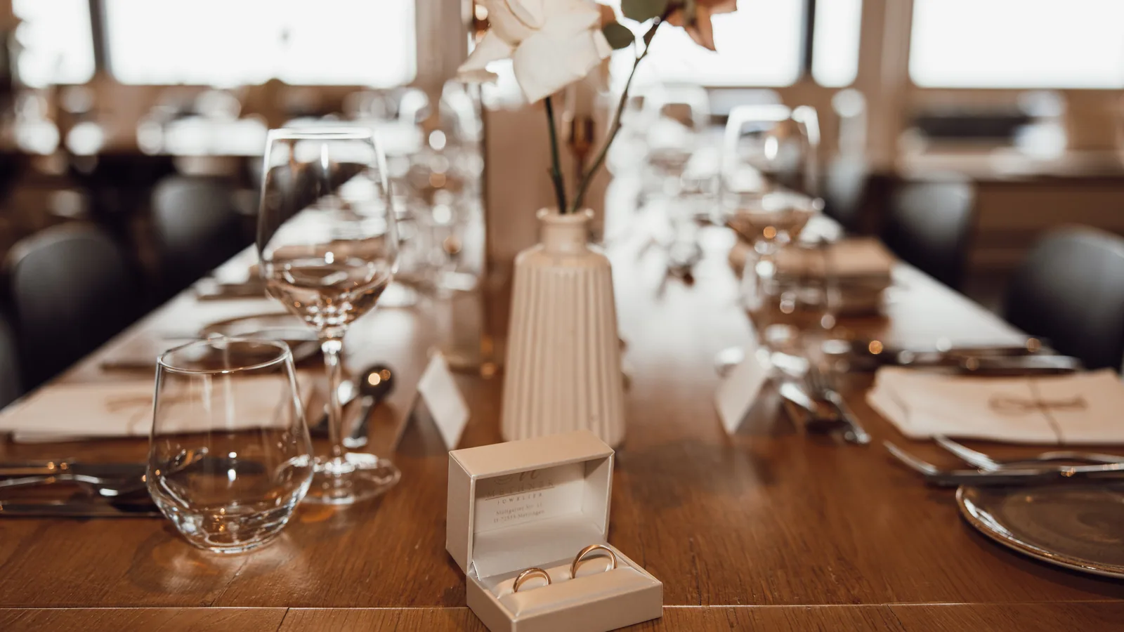 Gedeckte Hochzeitstafel im Gegenlicht: Ringschatulle im Vordergrund, dahinter Vase mit Rose und die Tischreihe in die Tiefe